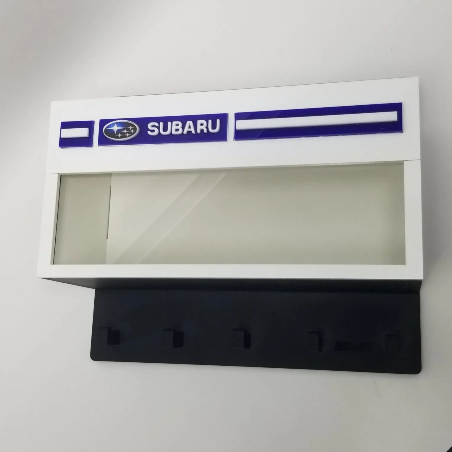 White rectangular Subaru wall key hook rack with glass display case and four black hooks.