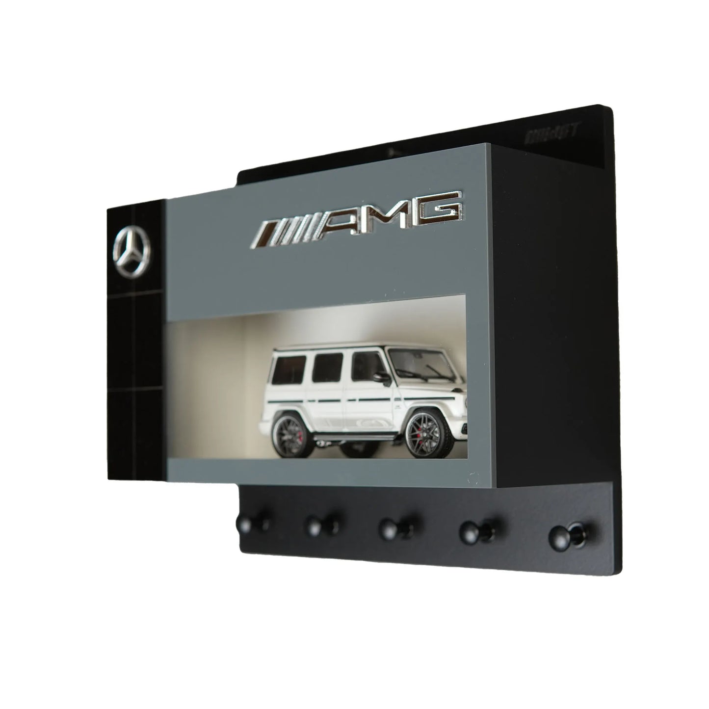Black Mercedes AMG wall-mounted key hook rack with a white scale model G-Class car inside a display case.