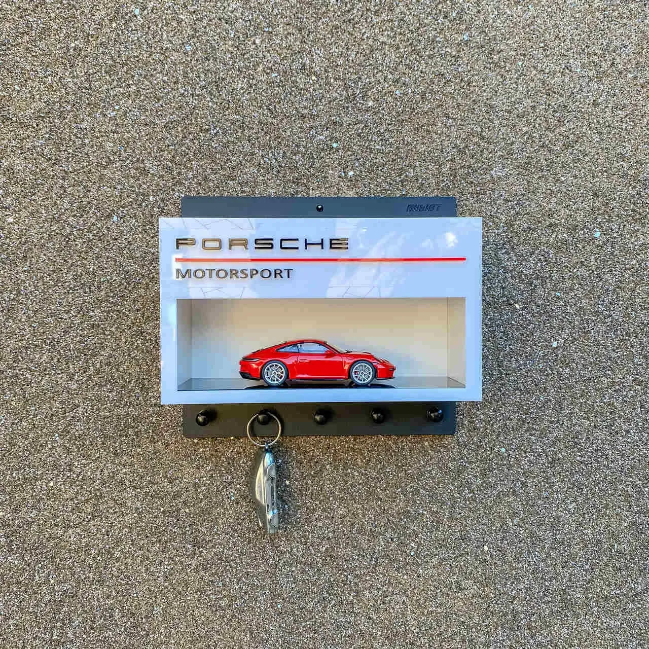 Gray Porsche Motorsport wall key hook rack with a red scale model car displayed inside and four black hooks below.