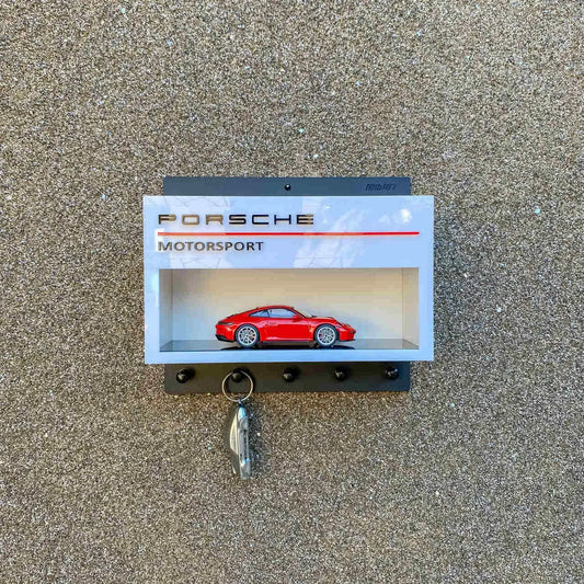 Gray Porsche Motorsport wall key hook rack with a red scale model car displayed inside and four black hooks below.