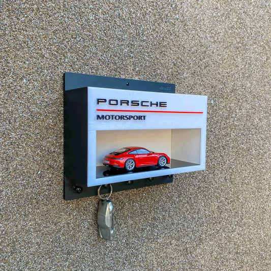 Black wall-mounted key hook rack with a white Porsche Motorsport scale model car display case and a hanging silver.