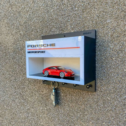 Wall-mounted Porsche Motorsport key hook rack with glass display case featuring red scale model car.