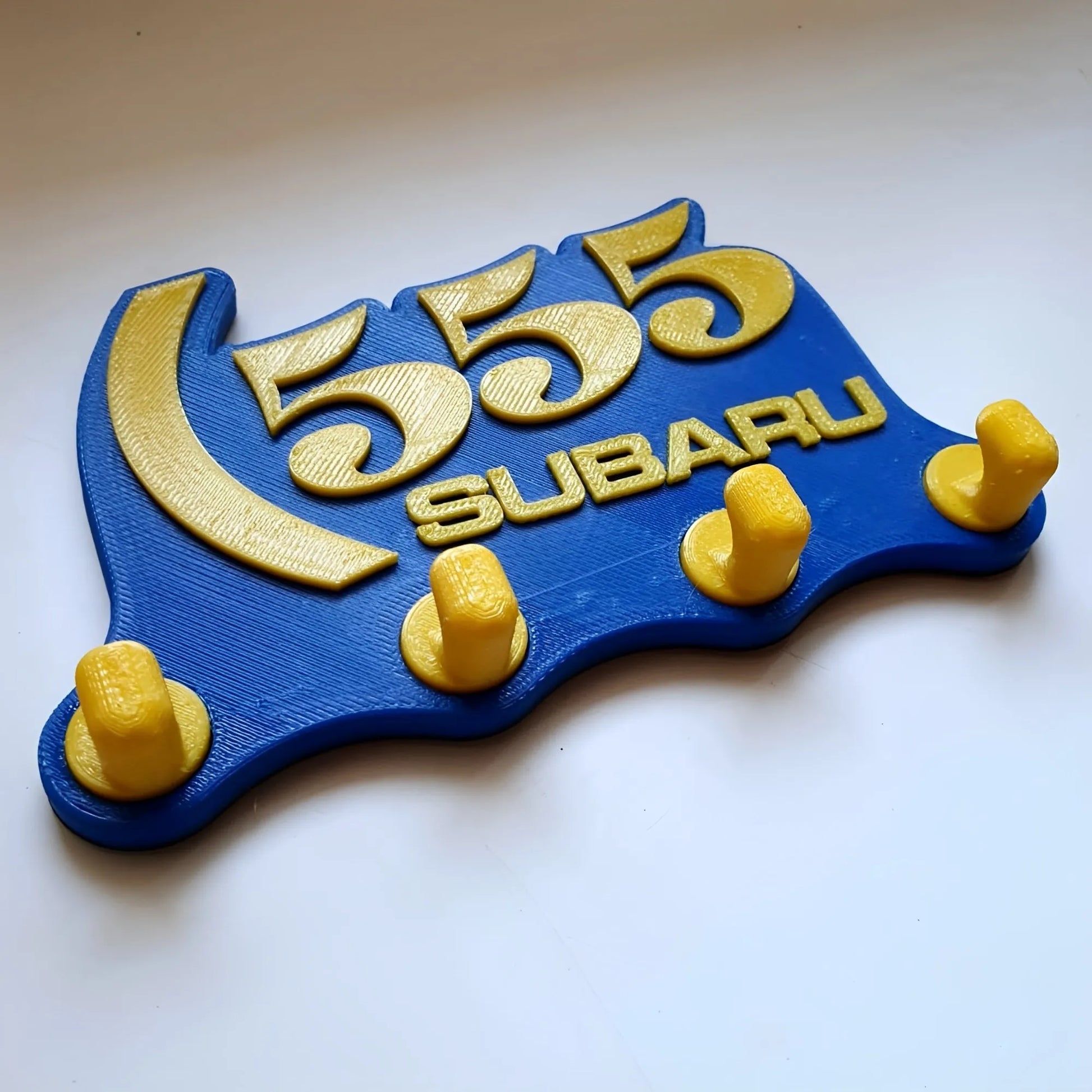 Blue and yellow Subaru 555 wall key hook rack with four yellow hooks and raised logo design.